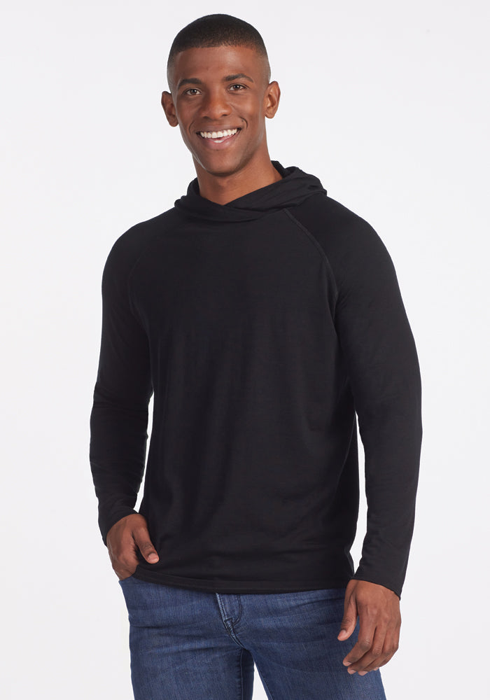 A person wearing a Base Camp Hoodie in black by Woolx and blue jeans smiles at the camera against a white background. 