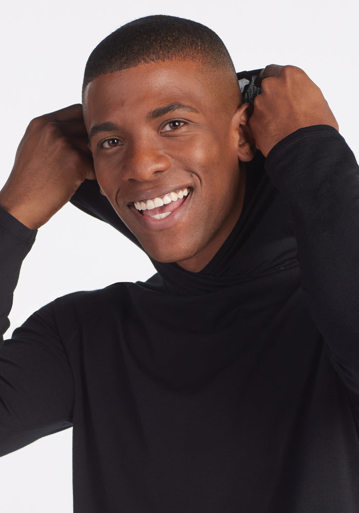 A person smiling while pulling up the hood of a Base Camp Hoodie - Black by Woolx, designed for outdoor wear, set against a plain white background.