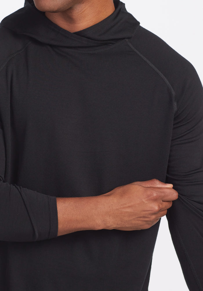 A person wearing a Woolx Base Camp Hoodie in black adjusts their sleeve, revealing a light-skinned arm. The hoodie showcases a smooth texture, making it ideal for outdoor clothing. The background is plain white.