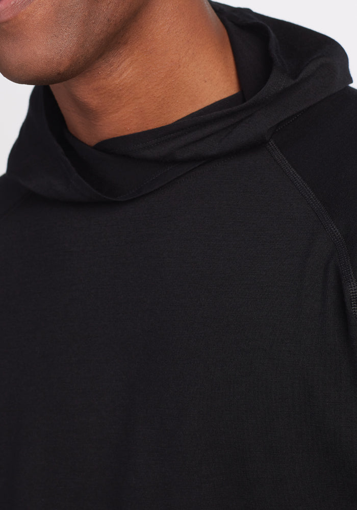 A person wearing the Base Camp Hoodie - Black from Woolx. Only part of the face and upper body is visible, with an emphasis on the Merino wool texture and hood detail. The background is plain white.