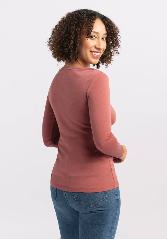 A person with curly hair smiles and looks over their shoulder, wearing the Woolx Kenzie Scoop Neck top in Brick Dust, featuring a stylish scoop neck design. Complemented by blue jeans, they stand gracefully against a plain white background.