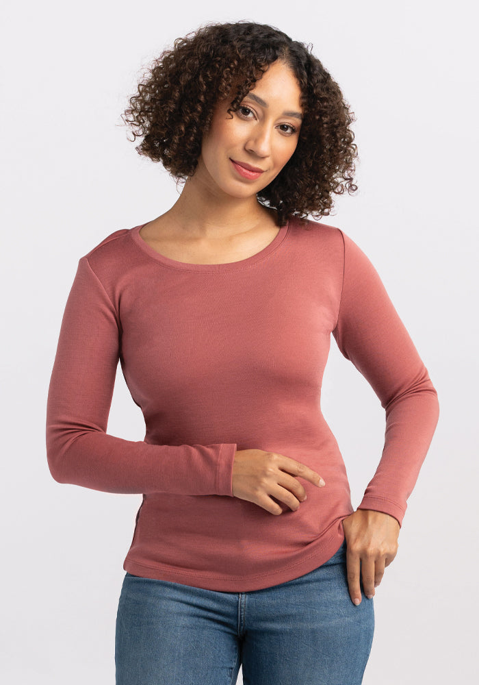 A person with curly hair wearing the Kenzie Scoop Neck in Brick Dust by Woolx and jeans stands against a plain white background. They look forward with a slight smile, one hand resting on their hip. 