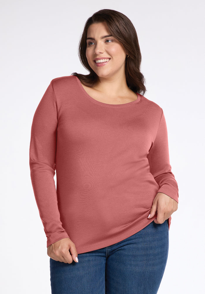 A woman with long dark hair smiles, wearing the Kenzie Scoop Neck in Brick Dust by Woolx, paired with blue jeans. She stands against a plain white background, exuding comfort and style in her moisture-wicking outfit. 