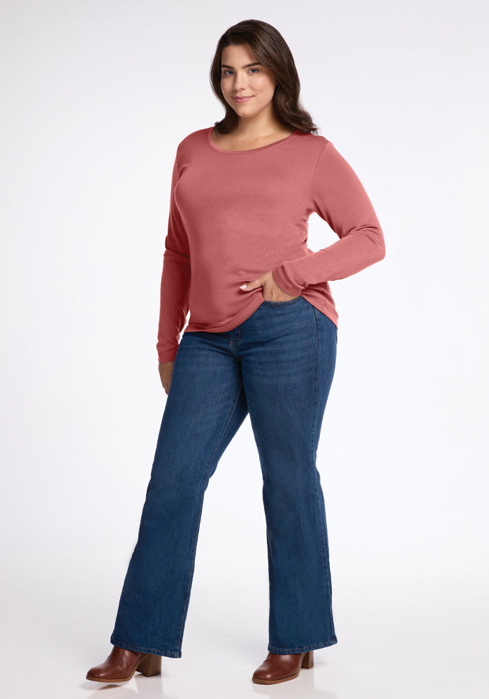 A person with long dark hair is posing confidently in a moisture-wicking Kenzie Scoop Neck top in Brick Dust from Woolx and blue flare jeans. One hand rests in their pocket, while the other is on their hip. Brown boots complete the casual look.