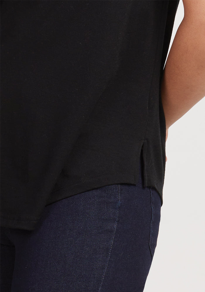 A person wearing the Bella Tunic Top in black from Woolx, paired with dark blue jeans, stands facing sideways, exuding warm-weather comfort. The top features a small side slit, and their left hand rests on their hip against the light gray backdrop.