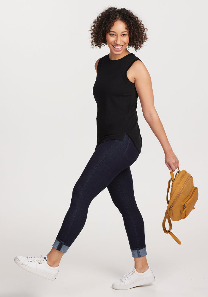 A person with curly hair smiles while walking in the Bella Tunic Top - Black by Woolx, paired with dark jeans and white sneakers, holding a yellow backpack. Embracing warm-weather comfort, they stroll against a plain, light-colored background.