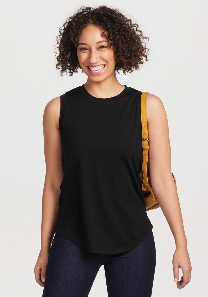 A person with curly hair smiles while wearing the sleeveless Bella Tunic Top in black by Woolx, paired with dark jeans. They carry a tan shoulder bag against a plain white background. 