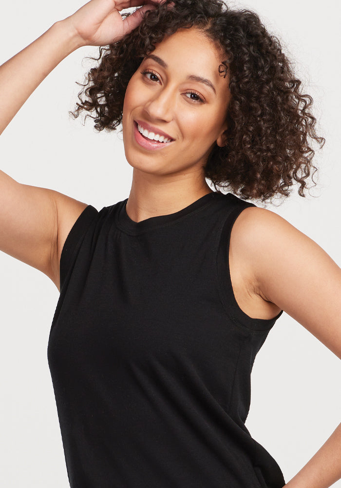 A smiling person with curly hair, wearing the Bella Tunic Top in black by Woolx, poses with one arm raised and hand gently touching their head, exuding warm-weather comfort.