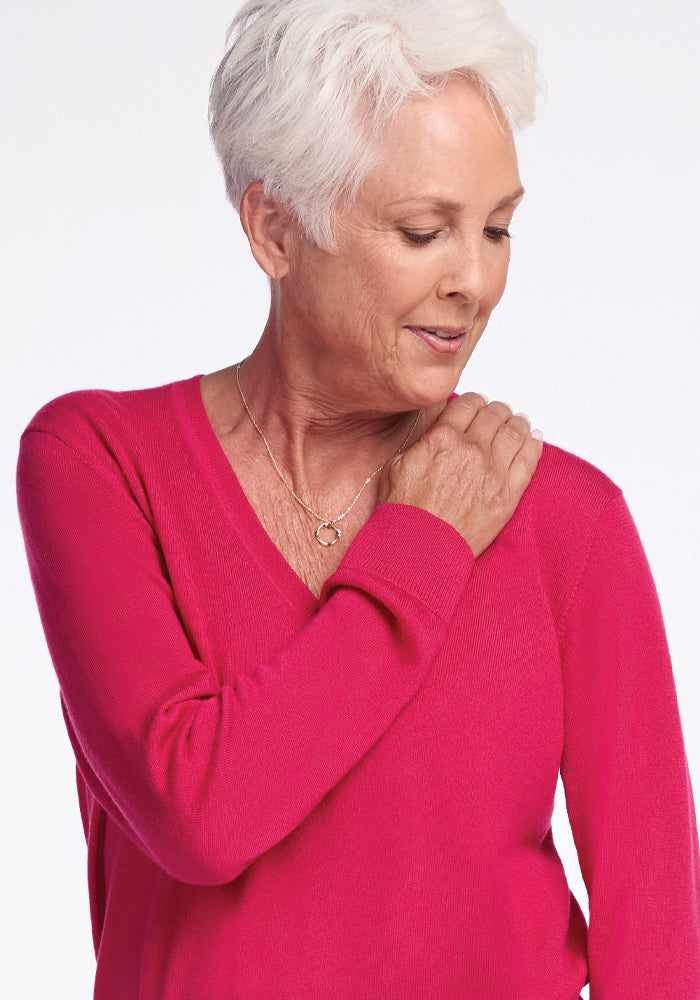 An elderly person with short white hair, wearing the Francesca Sweater in Berry Burst by Woolx and a delicate necklace, touches their shoulder. The person smiles gently while looking downwards against a plain white background.