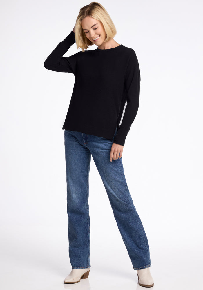 A person with short blonde hair wearing the Woolx Ainsley Sweater in black, featuring ribbed detailing, blue jeans, and white shoes is smiling while holding their head with one hand. They are standing against a plain white background.