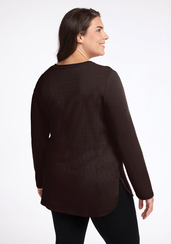 A person with long dark hair and a smiling expression is wearing the Blair Tunic in French Roast from Woolx, crafted from odor-resistant merino wool, accompanied by black pants. They are positioned against a plain white background, turned slightly to reveal both a side and back view.