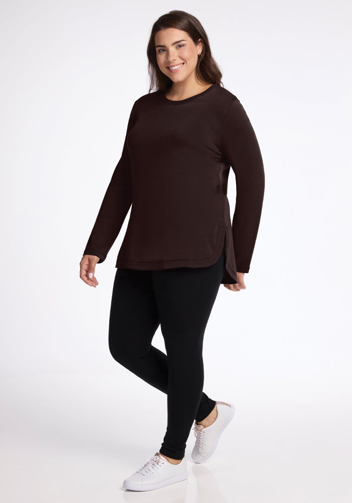 A woman is smiling in a Blair Tunic in French Roast by Woolx and black leggings. She stands against a plain white background, wearing odor-resistant sneakers designed for comfort and style.