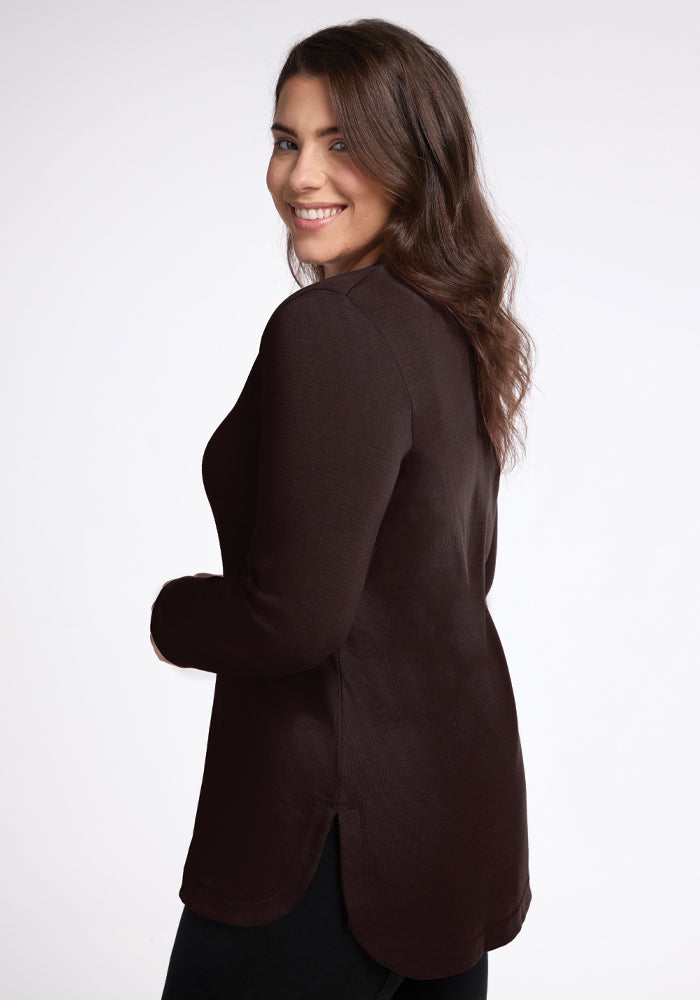 A person with long brown hair, wearing the Blair Tunic in French Roast by Woolx, is smiling and looking over their shoulder against a plain, light-colored background.