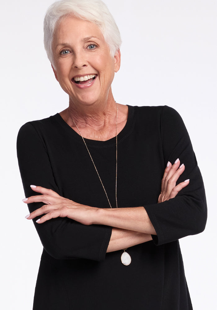 An older woman with short white hair smiles warmly, her arms crossed over a versatile Rissa A Line Dress by Woolx. She stands against a plain light background, exuding effortless style with her black dress and long necklace.