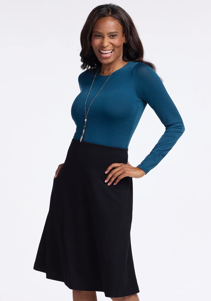 A woman smiles confidently with one hand on her hip, wearing a teal long-sleeve top paired with the stylish Marilyn Flowy Midi Skirt in Black from Woolx. Her ensemble is completed by a long necklace, all set against a plain white background.