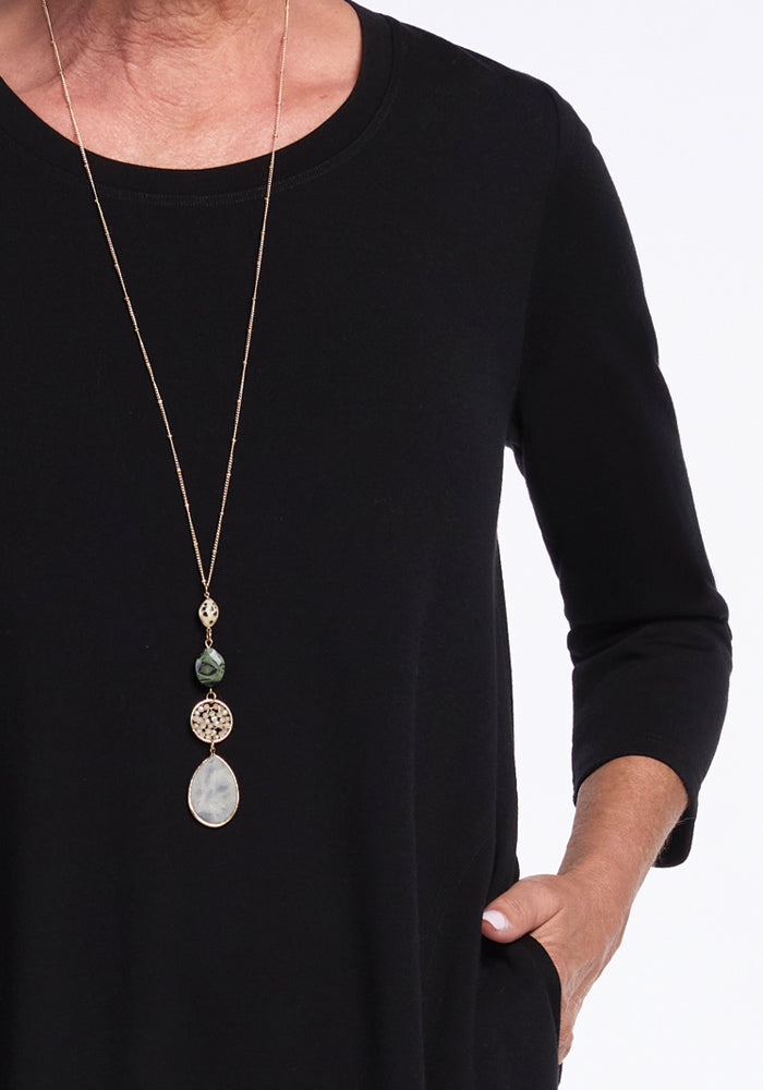 A person wearing the Rissa A Line Dress in black from Woolx, layering it effortlessly with a long necklace adorned with green and pale stones and a large teardrop-shaped pendant, stands against a white background with one hand in their pocket.