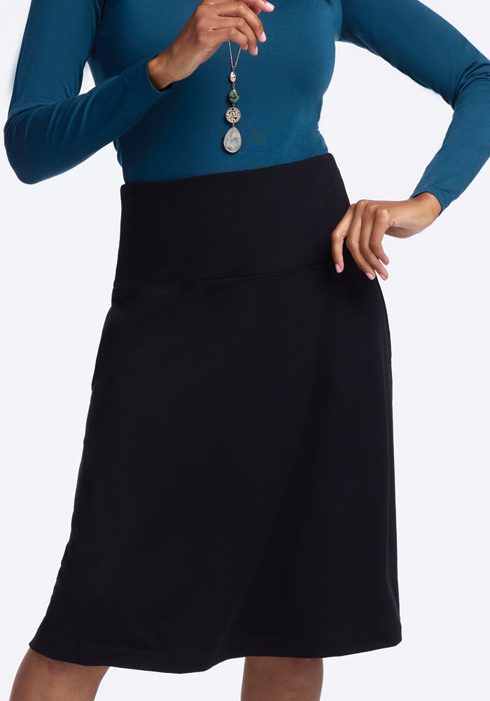 A person in a blue long-sleeve top and the Marilyn Flowy Midi Skirt - Black by Woolx, crafted from Merino wool with a wide waistband, holds a pendant necklace against a plain white backdrop.