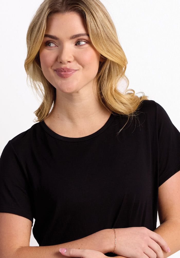 A person with long, wavy blond hair is smiling and looking to the side, highlighting their plain black Desi Nightgown made by Woolx. They accessorize with a delicate bracelet on their wrist, standing against a plain white wall.