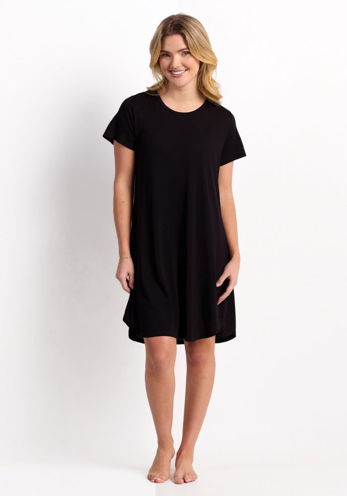 A person with long hair stands barefoot against a plain background, wearing the Desi Nightgown - Black by Woolx. They are smiling with hands relaxed by their sides, enjoying the temperature-regulating and odor-resistant qualities of the Feathertouch® Merino Wool fabric.