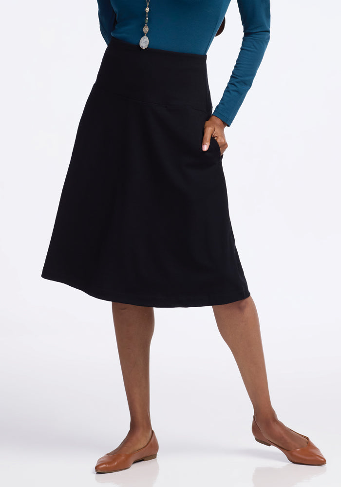 A person wearing a blue long-sleeve shirt, the Woolx Marilyn Flowy Midi Skirt in black, and brown flats stands with one leg slightly bent. They accessorize with a pendant necklace, set against a plain white background.