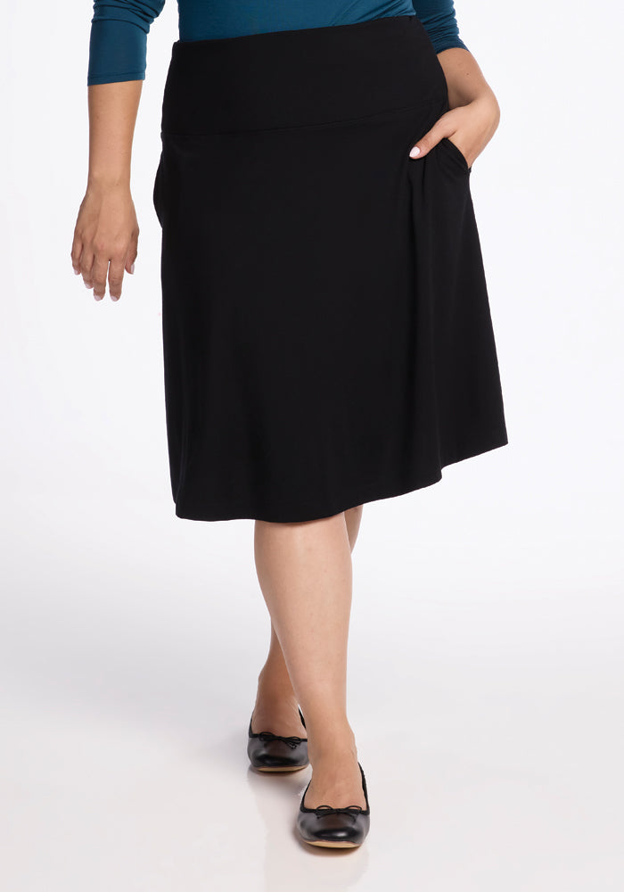 A person wearing the Marilyn Flowy Midi Skirt in Black from Woolx and a teal top, accompanied by black flats, strides forward effortlessly. Their hand rests in one pocket of the skirt’s flowy design, set against a plain white backdrop.