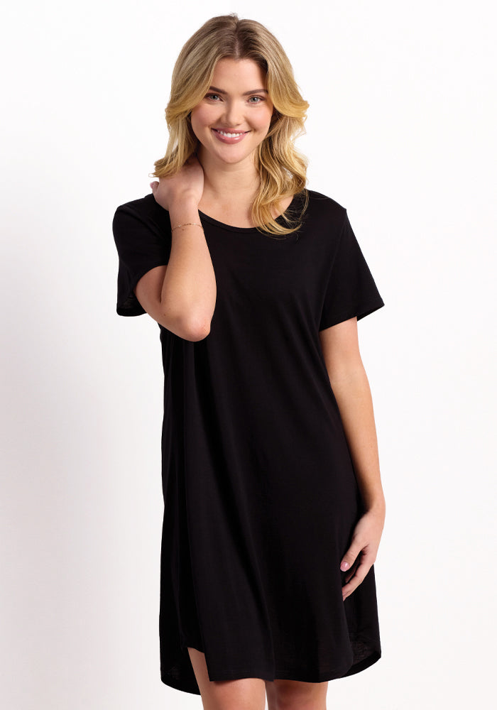 A person with blonde hair smiles while wearing the Desi Nightgown - Black from Woolx, made from Feathertouch® Merino Wool, which is celebrated for its temperature-regulating and odor-resistant qualities. They stand in a relaxed pose against a plain white background. 