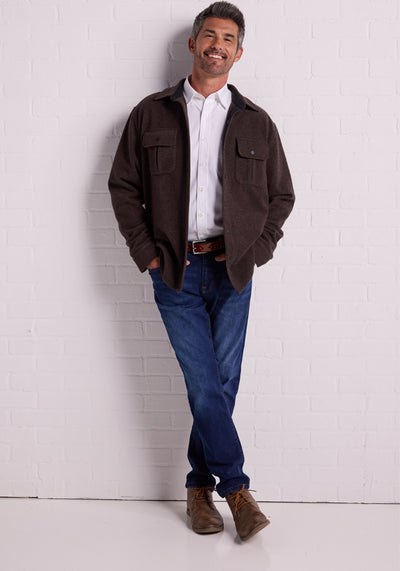 A person with short hair is standing against a plain background, wearing a Carbon Black Wilder Shirt Jac by Woolx over a Merino wool shirt and blue jeans. They are looking at the camera with a neutral expression and have their hands positioned near the front of their jacket, showcasing an ultra-functional look. | Trell is 6’2”, wearing a size M