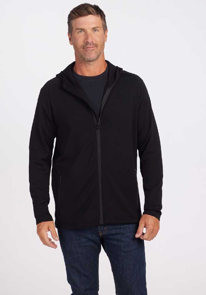 A man with short hair and a beard is standing against a plain white background. He is wearing the Boulder Hoodie - Black from Woolx over a dark t-shirt and blue jeans. The man has a neutral expression, and his hands are relaxed at his sides, embodying a casual style. 