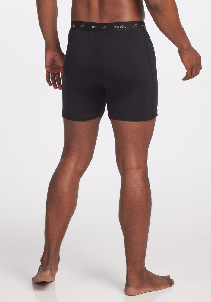 On a white background, a person stands barefoot wearing the Breathe Boxer Shorts in black from Woolx. Made from Australian Merino wool, these shorts ensure breathability and comfort. The image captures the figure from the back, emphasizing muscular legs and a naturally relaxed posture, with the waistband prominently featuring the "Woolx" label.