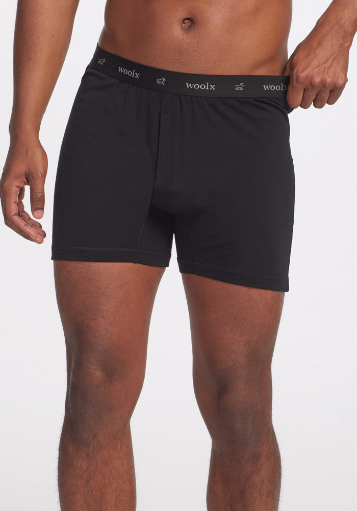 A person in black Breathe Boxer Shorts by Woolx, made from breathable Australian Merino wool, stands against a white background. The waistband prominently features the "woolx" brand name in white lettering. The individual is displayed from mid-thigh to waist, with one hand resting at their side.