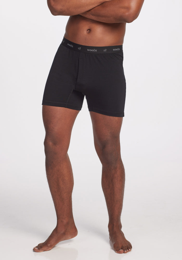 With arms crossed, a person models the Breathe Boxer Shorts in black by Woolx, featuring the brand name prominently on the waistband. Crafted from premium Australian Merino Wool, these breathable shorts ensure outstanding comfort against a plain white backdrop.