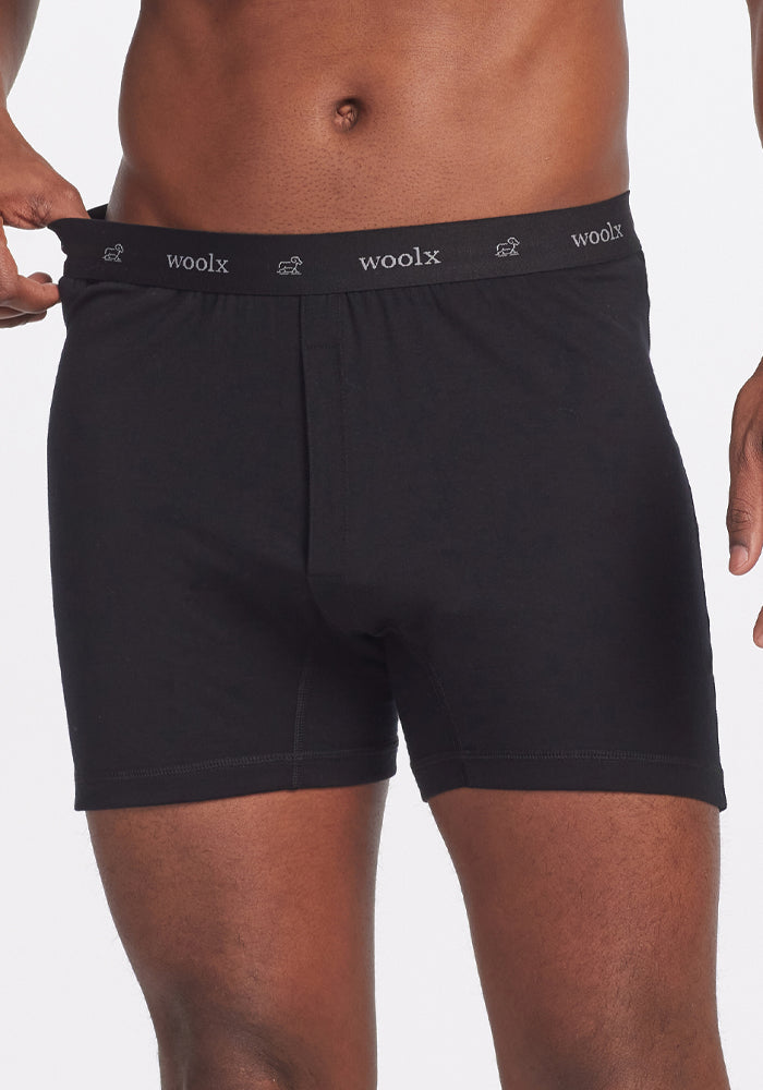 The individual is adjusting their black Breathe Boxer Shorts, which feature "Woolx" on the waistband, for comfort. These breathable boxer shorts are crafted from soft Australian Merino wool and offer all-day freshness. The image highlights the fine detail of the fabric from waist to mid-thigh.