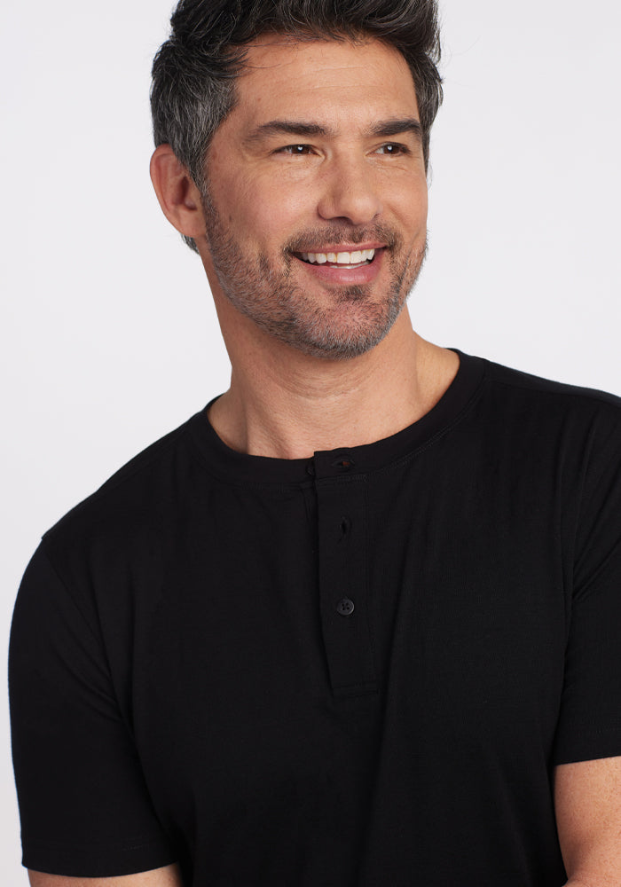 A smiling man with dark hair and a beard wears a black Bronx Henley shirt from Woolx. He is looking slightly to the side against a plain white background, exuding warmth and style with his Woolmark Certified choice.