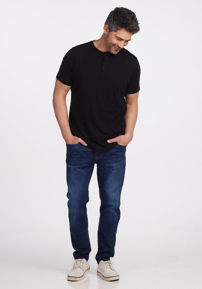 A person with short hair is wearing the Bronx Henley in black from Woolx, made from Merino wool and certified by Woolmark, paired with blue jeans. They are looking down and smiling while sporting white sneakers against a plain white background.