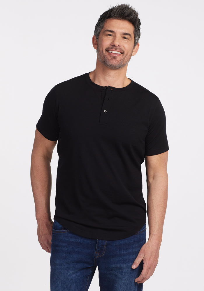 A smiling man with short, gray-streaked hair wears a black Bronx Henley from Woolx and blue jeans. He stands against a plain white background with one hand in his pocket, exuding casual elegance. 