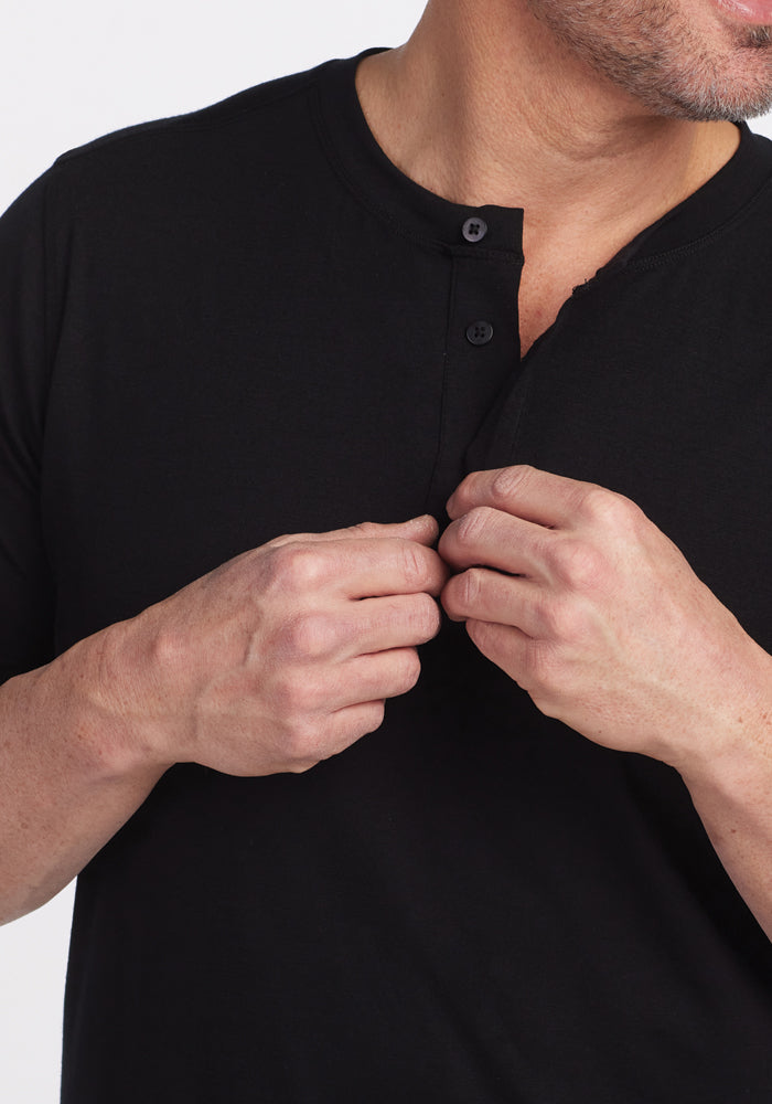 A person wearing the Woolx Bronx Henley in black, made from Merino wool, is shown from neck to waist. They are adjusting the top button, revealing a hint of their neck. The plain white background highlights the Woolmark Certified quality of the shirt.