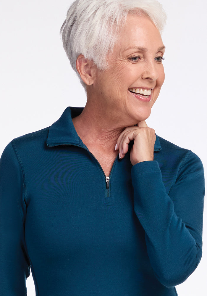 A smiling older woman with short white hair is pictured wearing a teal Brooke 1/4 Zip sweater by Woolx. She has one hand placed gently on her neck, looking slightly to the side, against a plain white background.