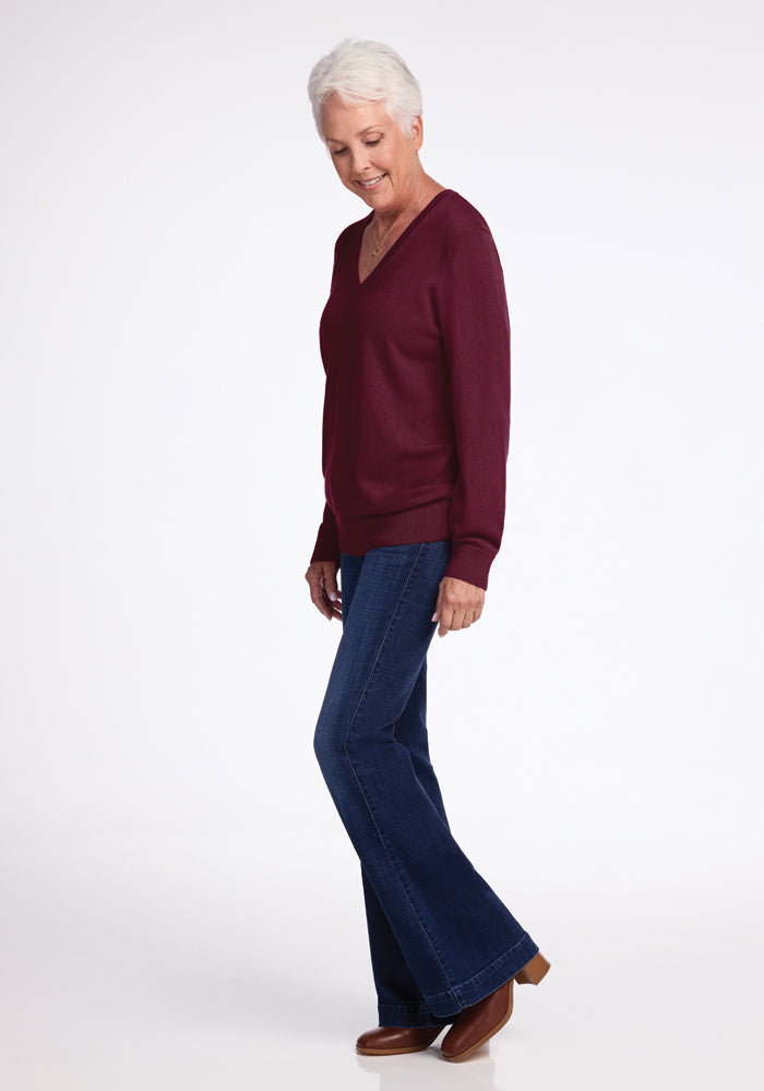 An older woman with short white hair is standing sideways, wearing a Woolx Francesca Sweater in Cabernet Crush, paired with blue jeans and brown shoes. She seems to be looking down against a plain white background.