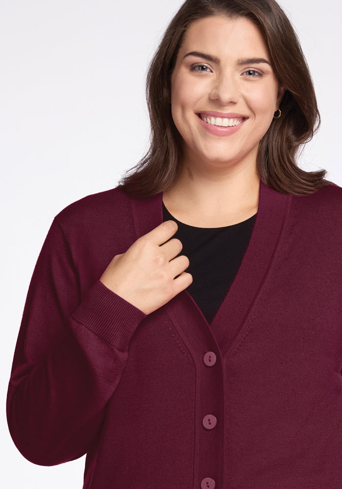 A person with shoulder-length brown hair smiles while wearing the Gia Sweater in Cabernet Crush from Woolx over a black top, featuring buttons down the front. The background is plain white.