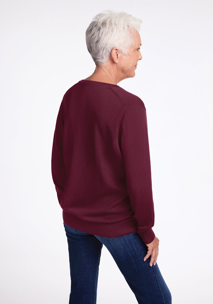 An older adult with short gray hair is standing and facing away, dressed in a Woolx Francesca Sweater in Cabernet Crush and blue jeans, against a plain white background.