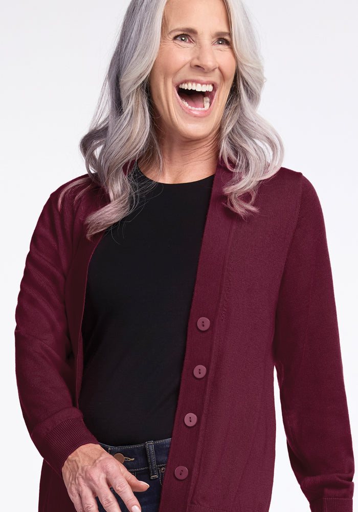 A woman with long gray hair is laughing. She is wearing a black shirt and Woolx's Gia Sweater in Cabernet Crush. The background is a plain white.