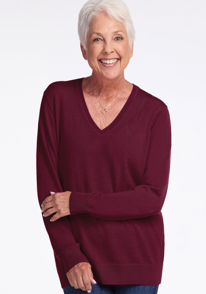 A smiling older woman with short white hair wears the Francesca Sweater in Cabernet Crush by Woolx and blue jeans. She stands against a plain white background with her arms relaxed.