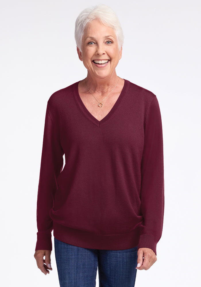 An older person with short white hair is smiling, wearing the Francesca Sweater in Cabernet Crush by Woolx and blue jeans, standing against a plain white background. 