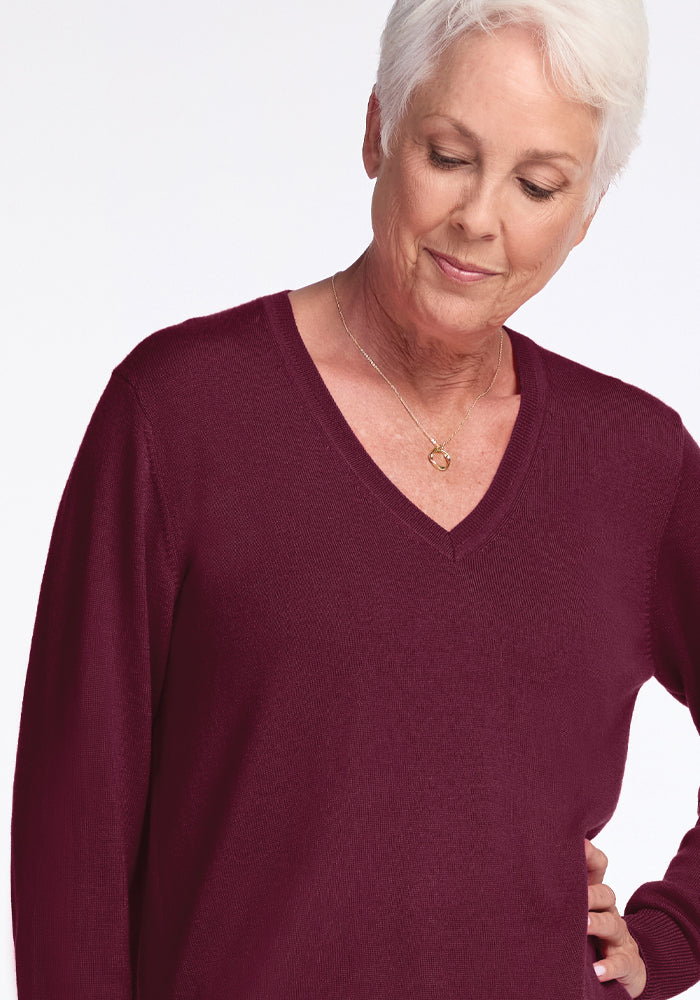 An older adult with short white hair is wearing a Woolx Francesca Sweater in Cabernet Crush and a necklace. They have a gentle expression and are looking downwards against a plain white background.