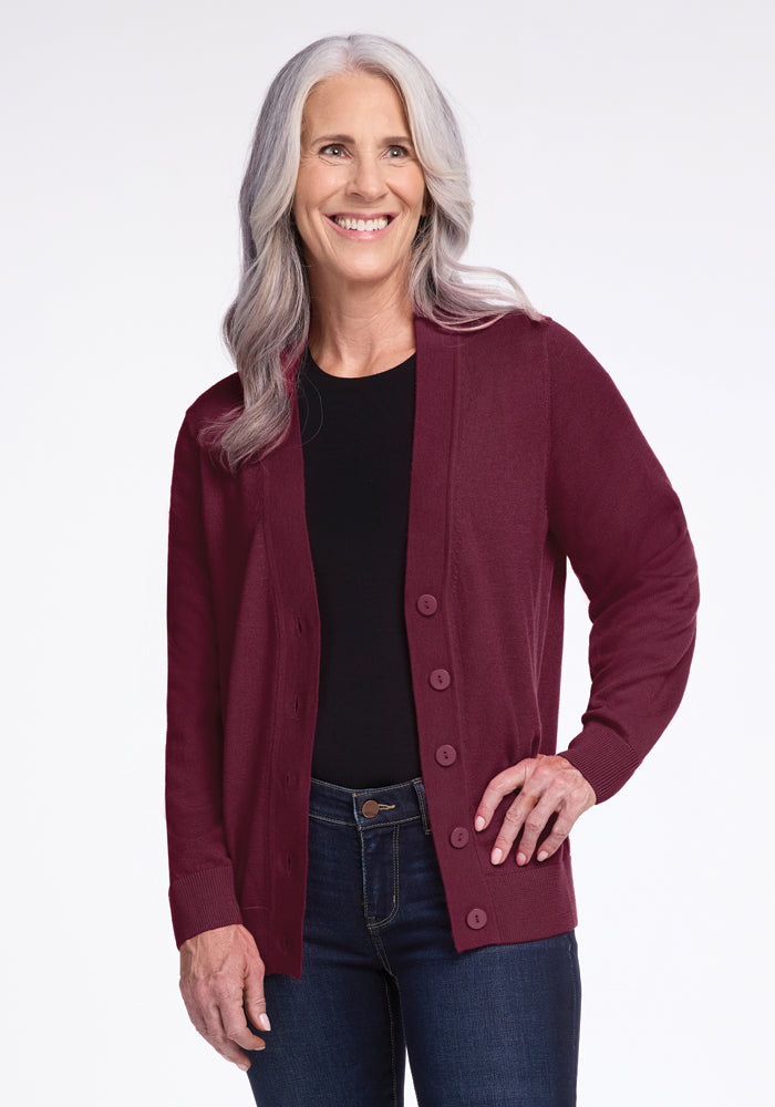 A smiling woman with long gray hair wears the Gia Sweater in Cabernet Crush by Woolx over a black shirt and blue jeans. She stands against a plain white background, with one hand on her hip. 