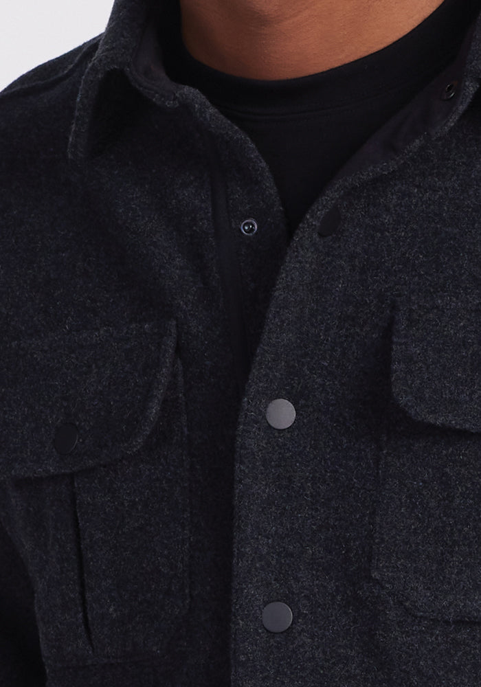 The image depicts an individual dressed in the Woolx Wilder Shirt Jac in Carbon Black, layered over a black shirt. This ultra-functional wool jacket, made from Merino wool, includes two chest pockets and a button-up design. The focus is on the upper half of the torso, with no facial features visible.