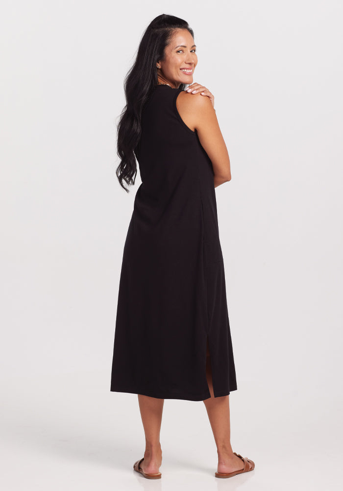 A woman with long dark hair smiles while slightly facing left, wearing the Cassie Dress - Black by Woolx. This sleeveless merino wool dress is ideal for travel and paired with sandals against a plain white background.