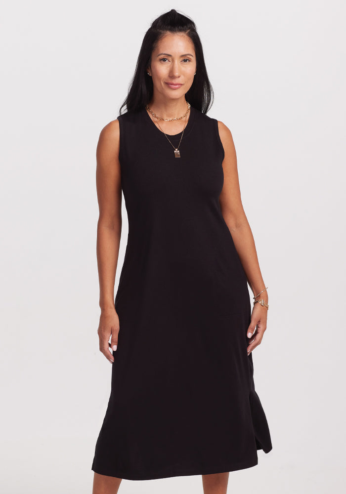 A woman with long dark hair wears the sleeveless Woolx Cassie Dress in black against a plain light background. Her subtle jewelry enhances the outfit as she looks forward with a slight smile. 