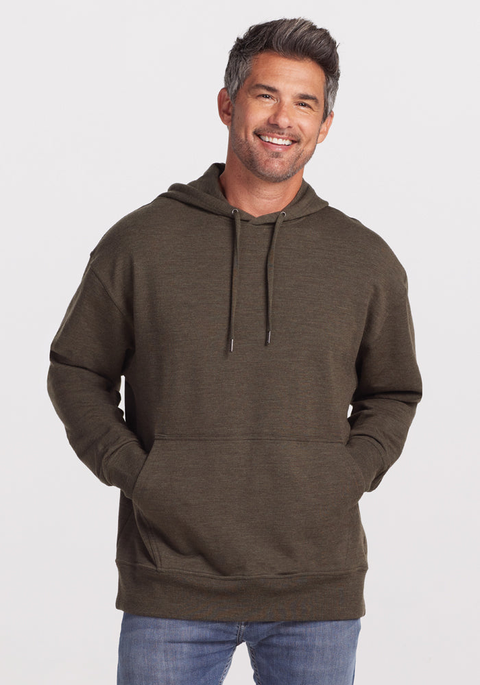 A man with short, graying hair and a beard is smiling and posing in the Woolx Chase Hoodie in Dark Moss. His hands are in the front pockets of the hoodie, and he is wearing blue jeans. The background is plain and light-colored. 