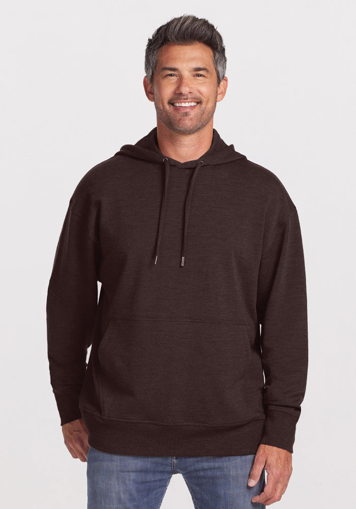 A smiling man with short, gray hair is wearing a Woolx Chase Hoodie in Maple Melange, made of Merino wool, along with blue jeans. He is standing against a plain white background, with his hands by his sides. The men's hoodie features a front pocket and drawstrings. 